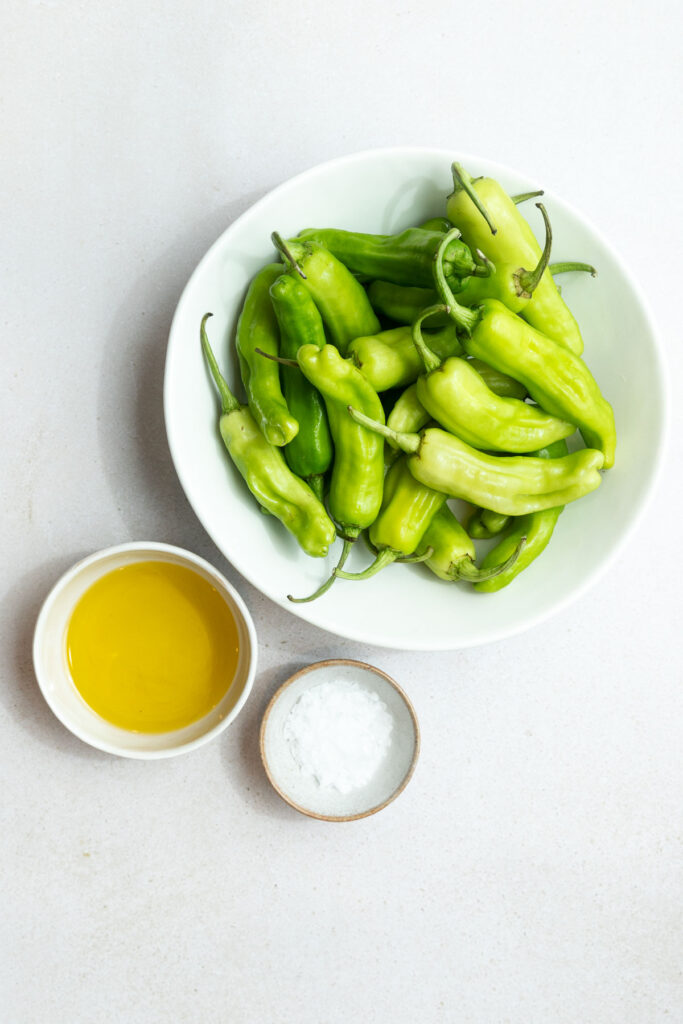 A white bowl filled with fresh green shishito peppers sits beside a small dish of olive oil and a small dish of coarse salt on a white surface, perfect for trying a simple shishito peppers recipe.