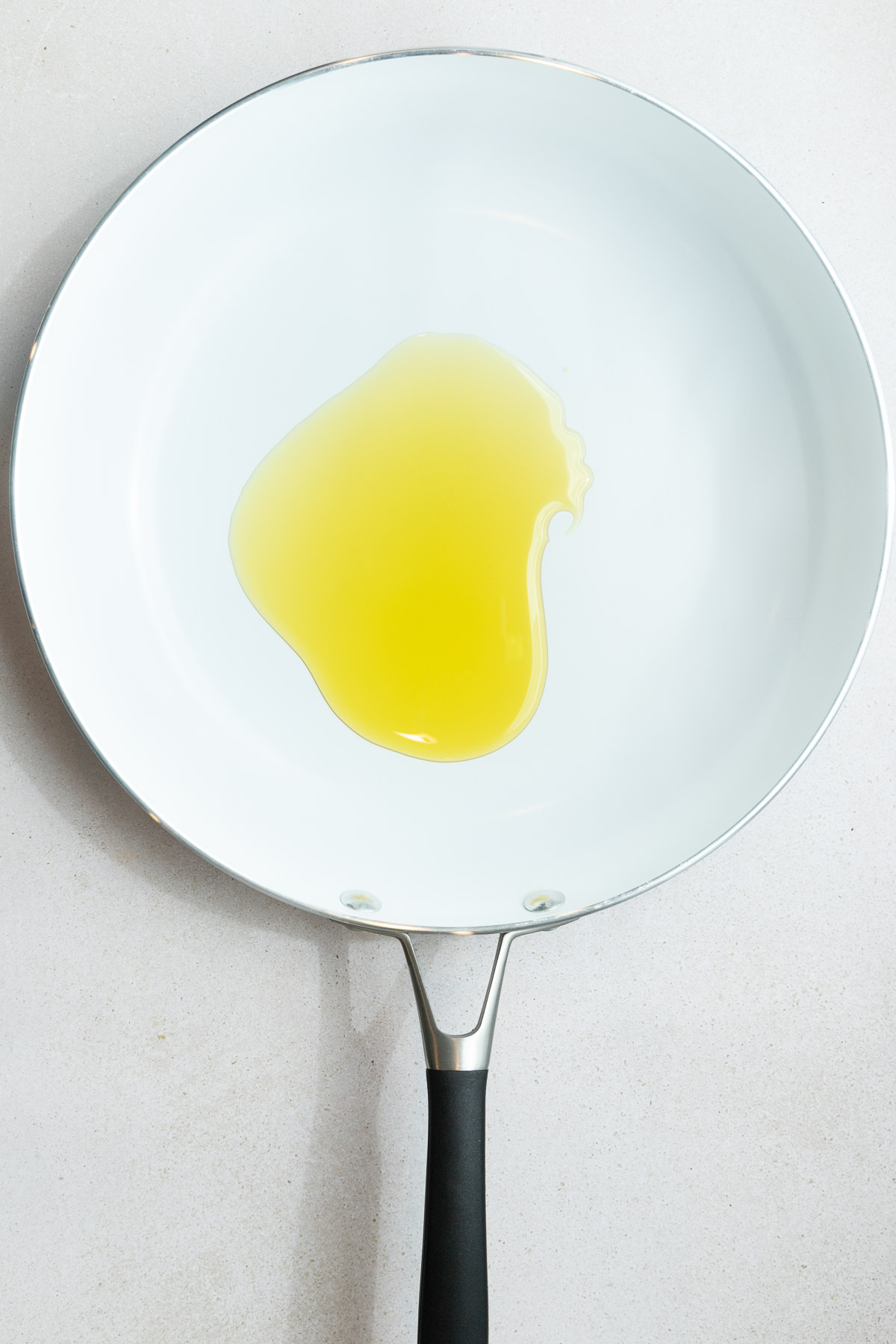 A white ceramic frying pan with a black handle contains a pool of yellow cooking oil, ready for how to cook shishito peppers, placed on a light-colored countertop.