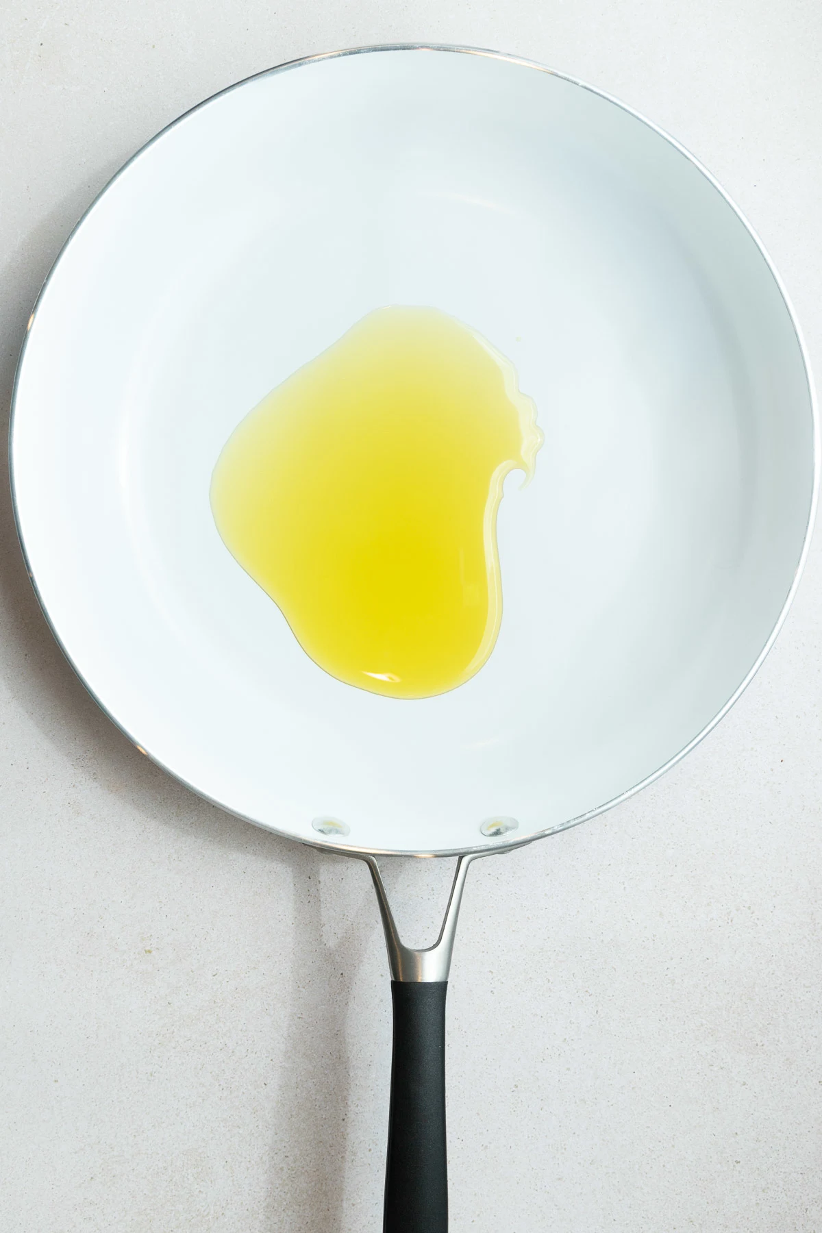 A white ceramic frying pan with a black handle contains a pool of yellow cooking oil, ready for how to cook shishito peppers, placed on a light-colored countertop.