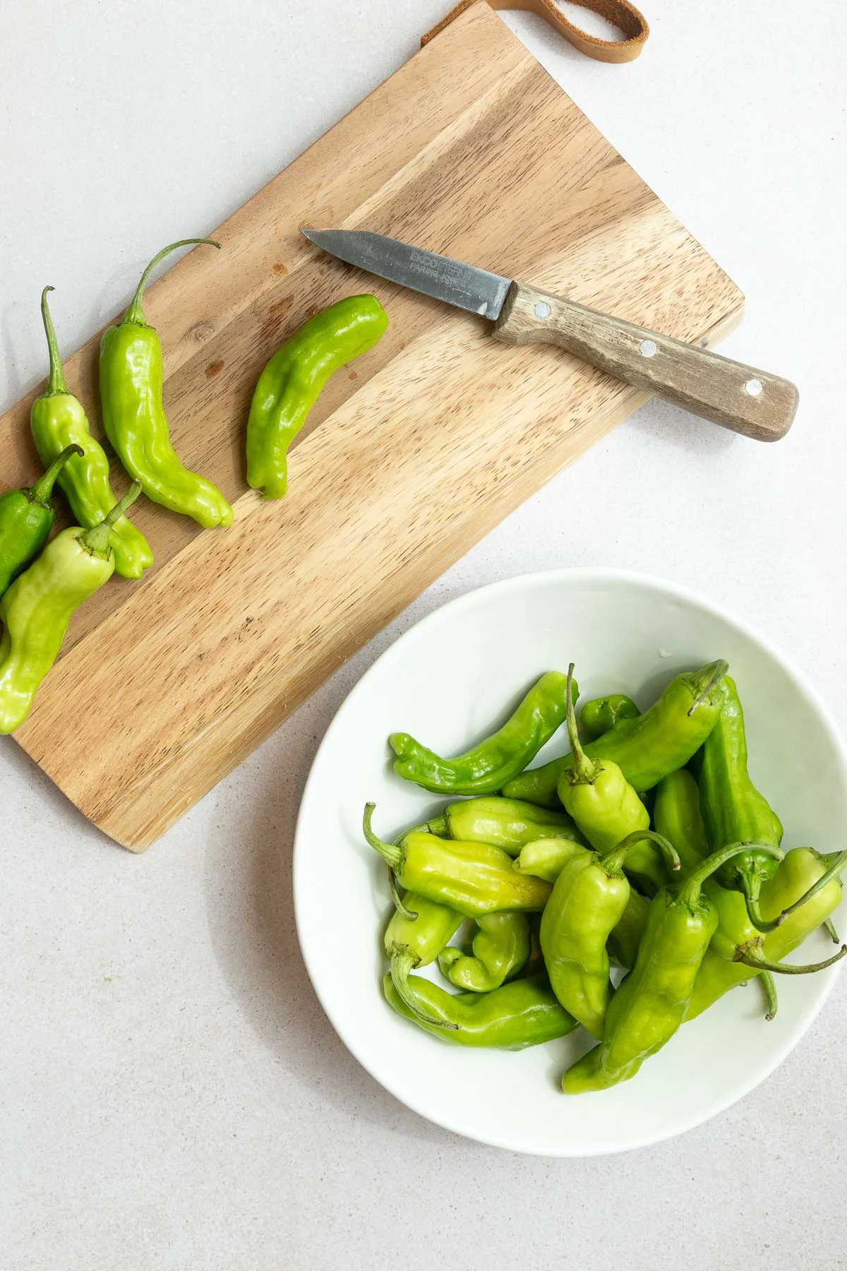 A wooden cutting board with a paring knife and several fresh green peppers, perfect for learning how to cook shishito peppers, sits next to a white bowl of more green peppers on a light countertop.