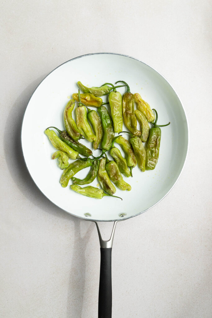 A white frying pan with a black handle containing several blistered shishito peppers arranged in a loose circle on a light-colored surface.