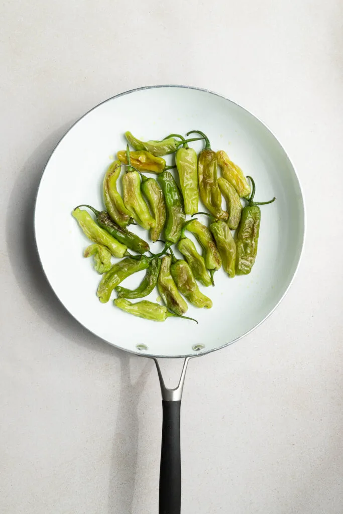A white frying pan with a black handle containing several blistered shishito peppers arranged in a loose circle on a light-colored surface.