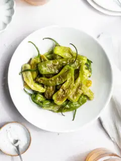 A white bowl filled with cooked green shishito peppers topped with flaky salt sits on a white surface next to a bowl of salt, a spoon, and partial plates—perfect for anyone learning how to cook shishito peppers.
