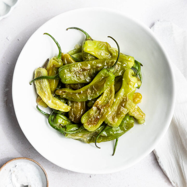 A white bowl filled with cooked green shishito peppers topped with flaky salt sits on a white surface next to a bowl of salt, a spoon, and partial plates—perfect for anyone learning how to cook shishito peppers.