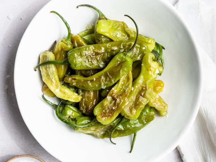 A white bowl filled with cooked green shishito peppers topped with flaky salt sits on a white surface next to a bowl of salt, a spoon, and partial plates—perfect for anyone learning how to cook shishito peppers.