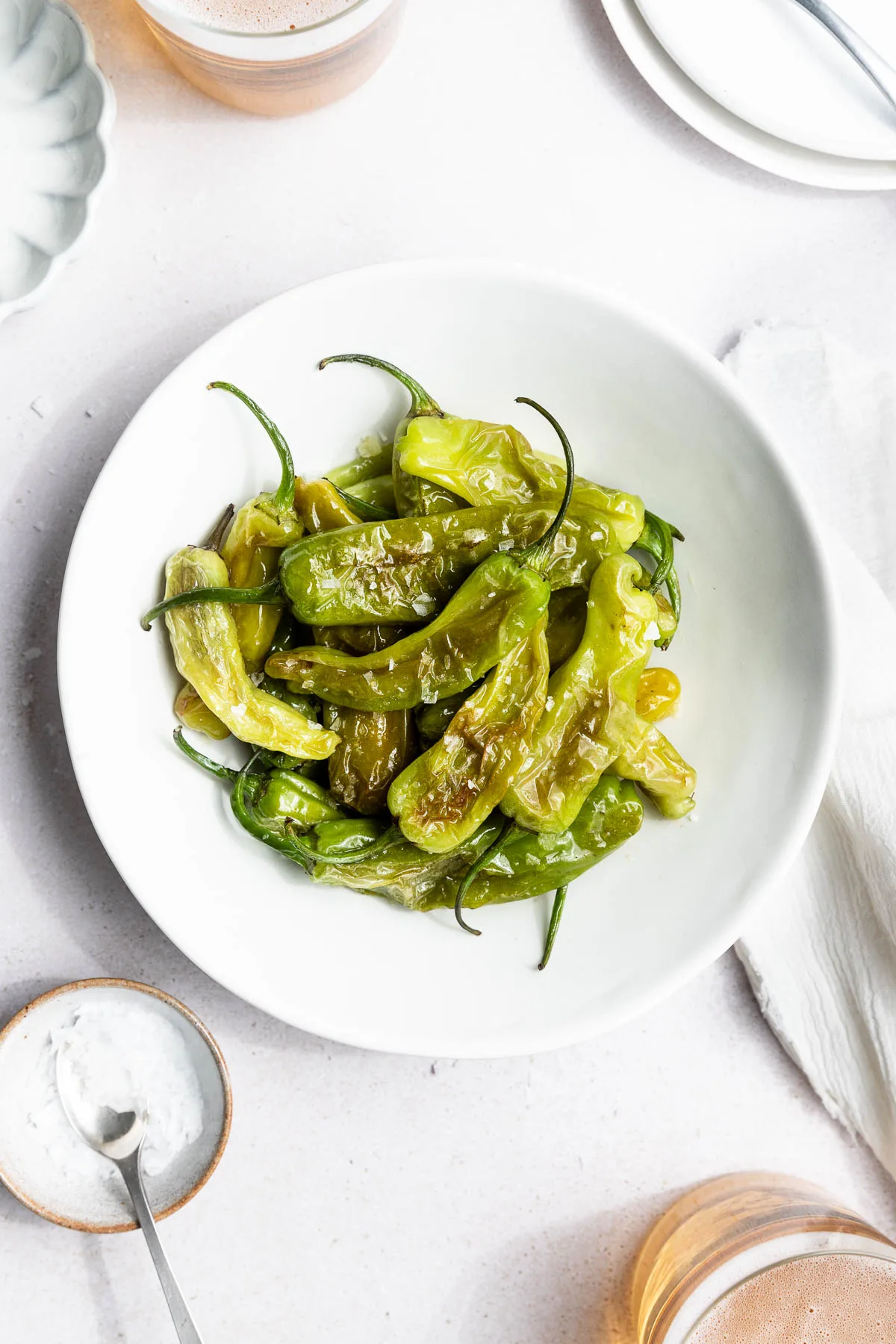 A white bowl filled with cooked green shishito peppers topped with flaky salt sits on a white surface next to a bowl of salt, a spoon, and partial plates—perfect for anyone learning how to cook shishito peppers.