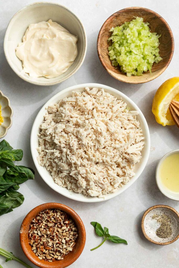 A bowl of shredded chicken meat surrounded by small bowls of mayonnaise, chopped celery, pecans, lemon, salt, pepper, lemon juice, and fresh basil—perfect for adding to your favorite lemon basil chicken salad.