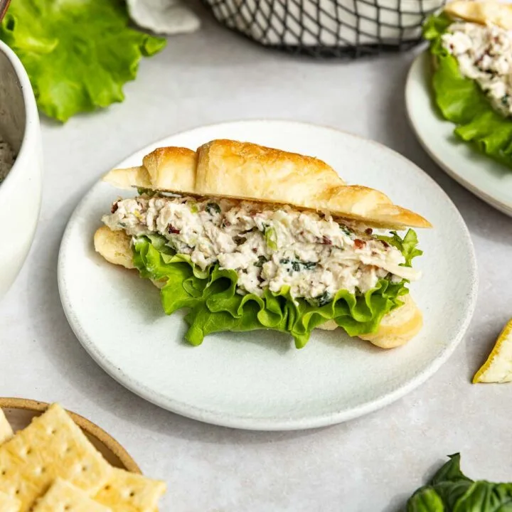 A croissant sandwich filled with copycat Lauryn's Lemon Basil chicken salad and leafy lettuce sits on a white plate, surrounded by crackers, fresh basil, and a basket of croissants in the background.
