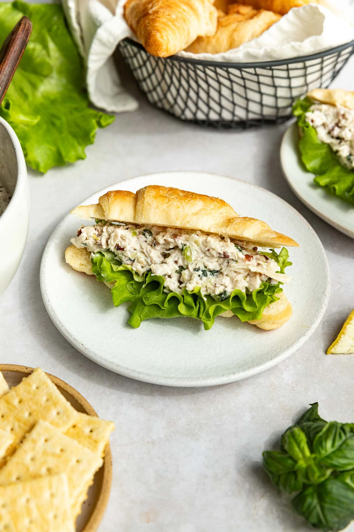 A croissant sandwich filled with copycat Lauryn's Lemon Basil chicken salad and leafy lettuce sits on a white plate, surrounded by crackers, fresh basil, and a basket of croissants in the background.