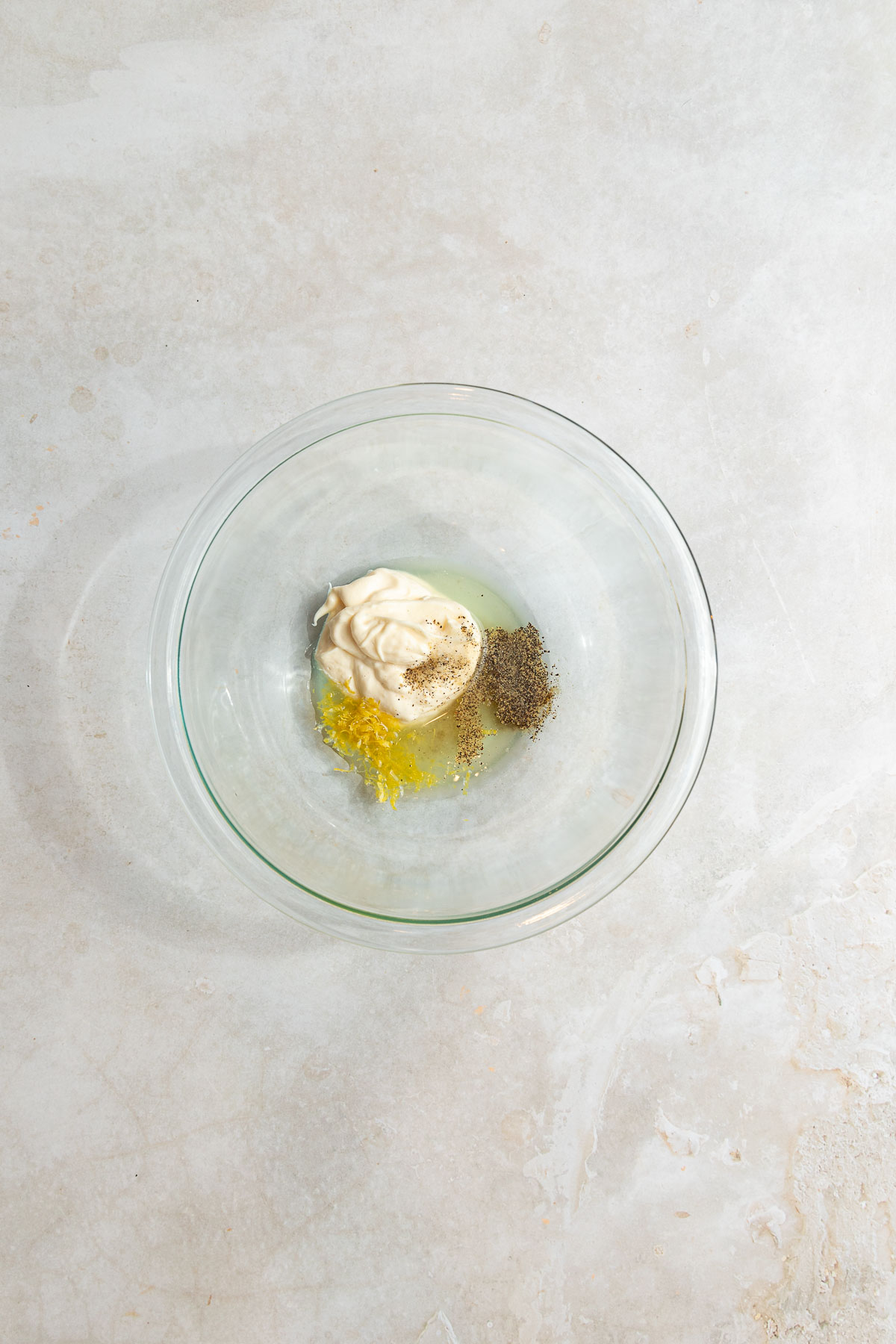 A clear glass bowl on a light countertop contains mayonnaise, black pepper, lemon zest, and a small amount of liquid, ready to be mixed for a fresh lemon basil chicken salad.