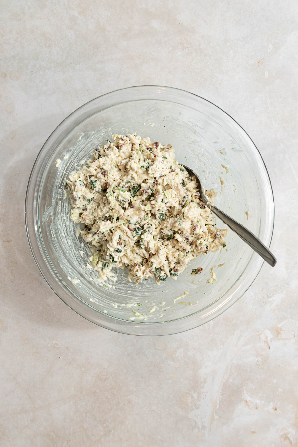 A glass mixing bowl containing a creamy lemon basil chicken salad mixture with chopped herbs and other ingredients, with a metal spoon resting inside. The bowl is placed on a light-colored countertop.