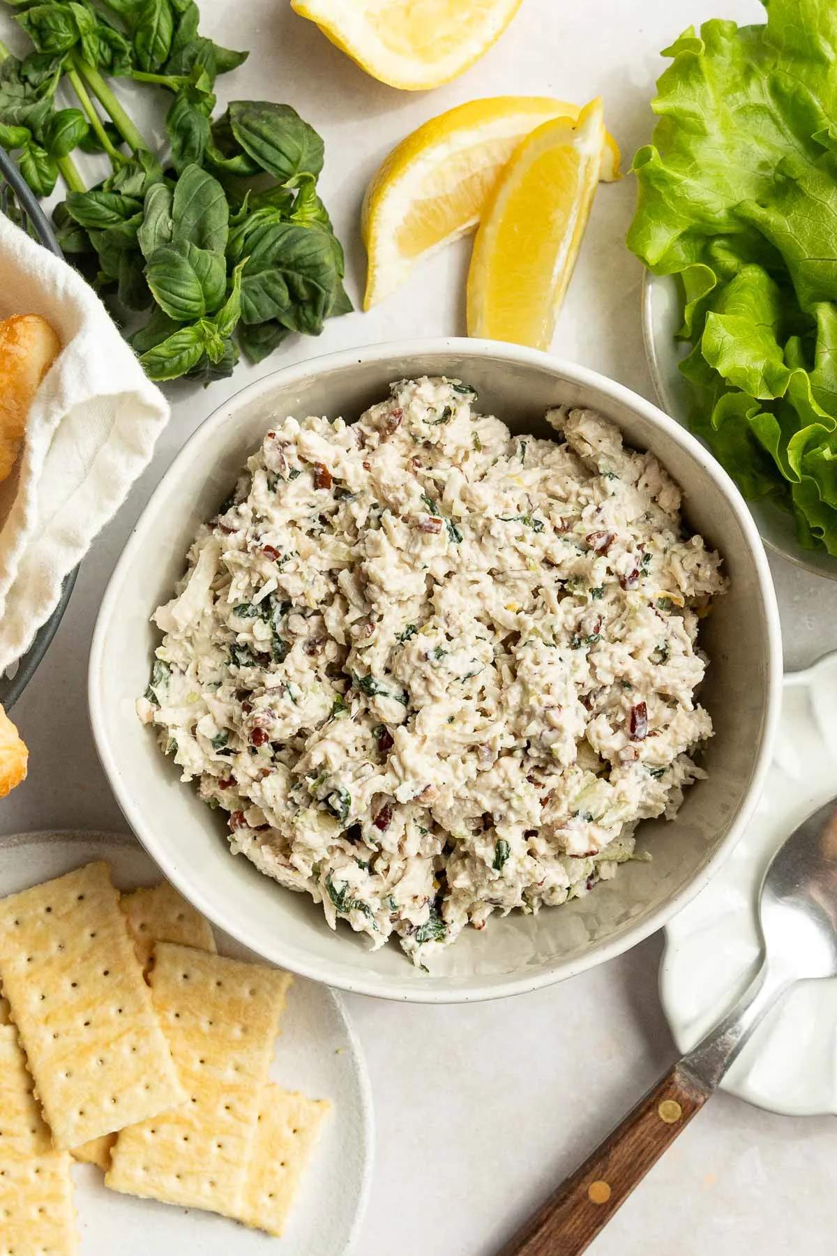A bowl of chicken salad with basil sits on a table, surrounded by lettuce leaves, lemon wedges, fresh herbs, saltine crackers, a serving spoon, and part of a bread basket.