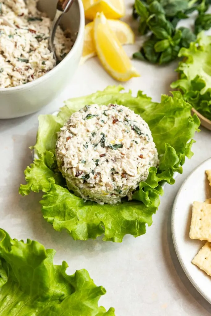 A serving of herbed chicken salad sits on a large green lettuce leaf, surrounded by lemon wedges, fresh herbs, a bowl of more chicken salad, and a plate with crackers.