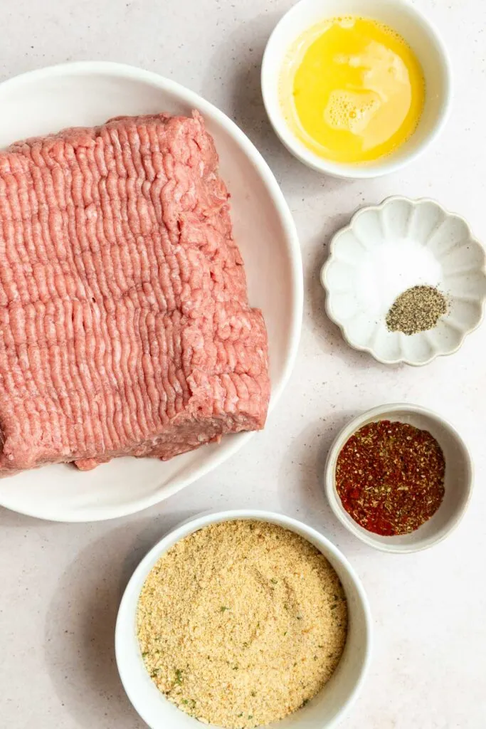 A plate of raw ground meat, perfect for a turkey meatballs recipe, is surrounded by small bowls of beaten egg, black pepper, red seasoning, and breadcrumbs on a light-colored surface.