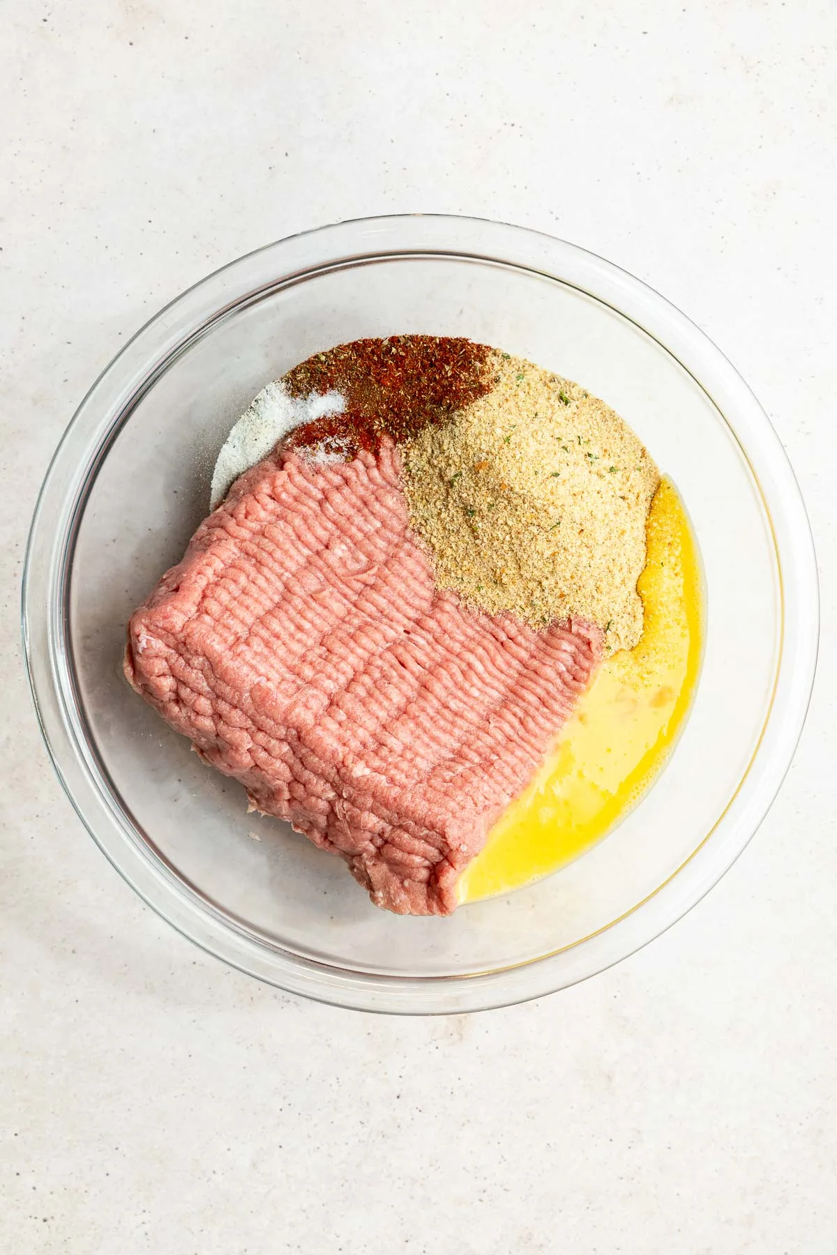 A glass bowl containing ground turkey meatball ingredients—ground meat, breadcrumbs, egg, and various spices—arranged in separate sections on a light-colored surface.