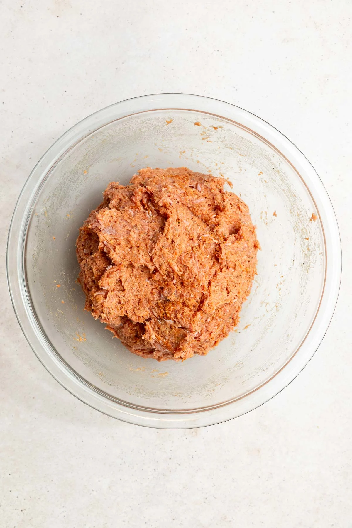 A glass mixing bowl filled with a blended mixture for baked turkey meatballs, made from ground meat and seasonings, sits on a light-colored countertop.