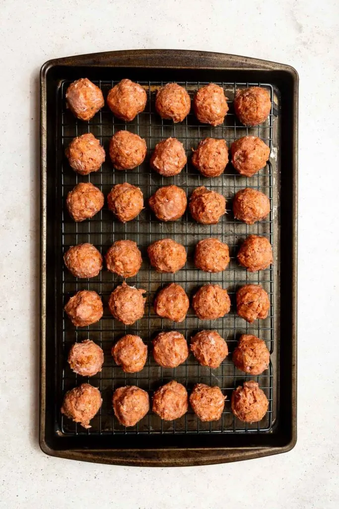 A baking tray lined with a wire rack holds five rows of uncooked turkey meatballs, evenly spaced and ready for the oven on a light surface.