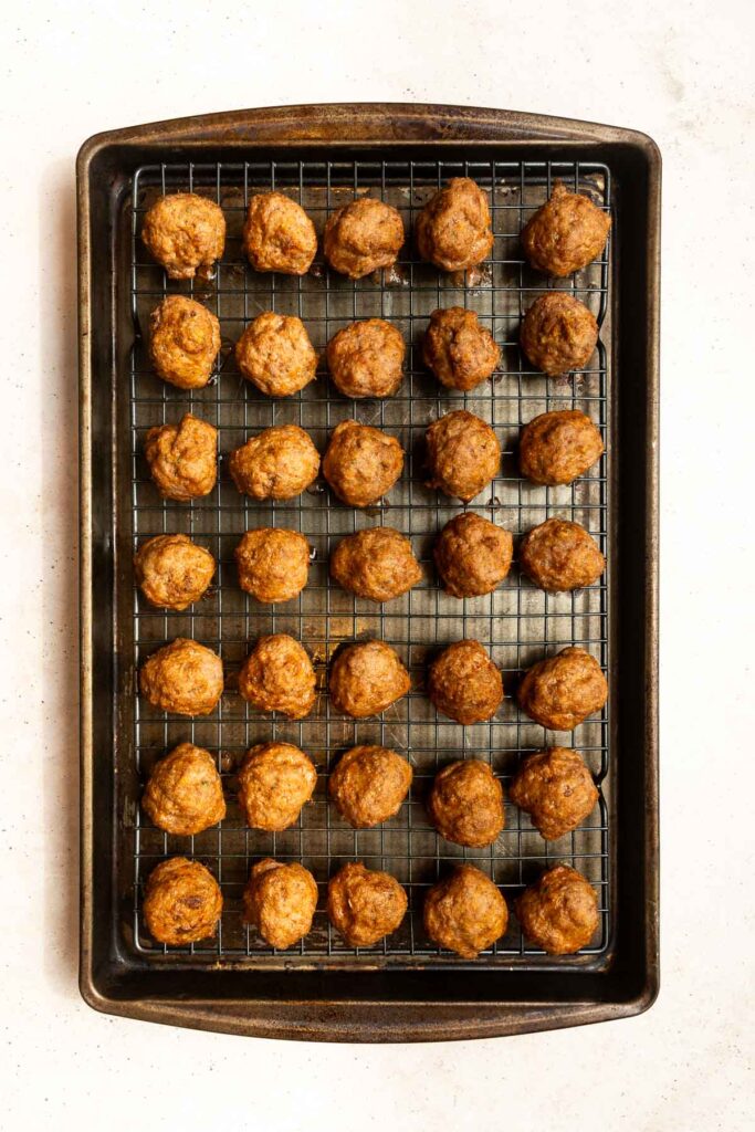 A baking tray lined with a wire rack holds five rows of evenly spaced, golden-brown Oven Baked Turkey Meatballs on a white background.