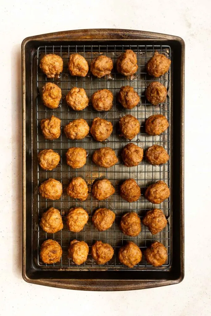 A baking tray lined with a wire rack holds five rows of evenly spaced, golden-brown Oven Baked Turkey Meatballs on a white background.