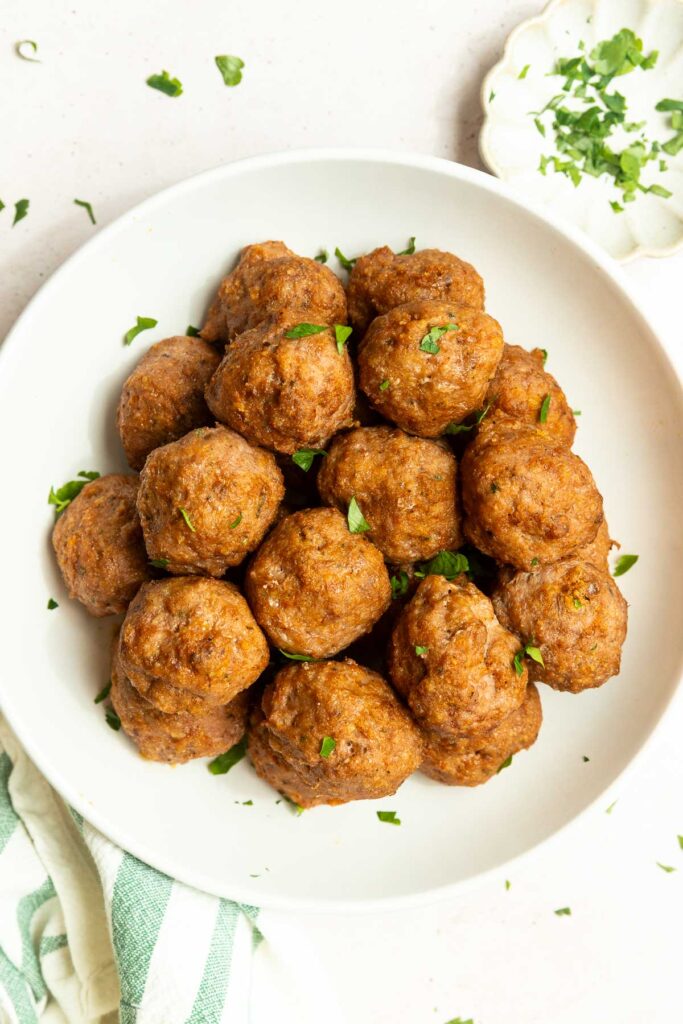 A white bowl filled with browned Oven Baked Turkey Meatballs, garnished with chopped fresh parsley, sits on a light surface next to a small dish of more parsley and a striped kitchen towel.