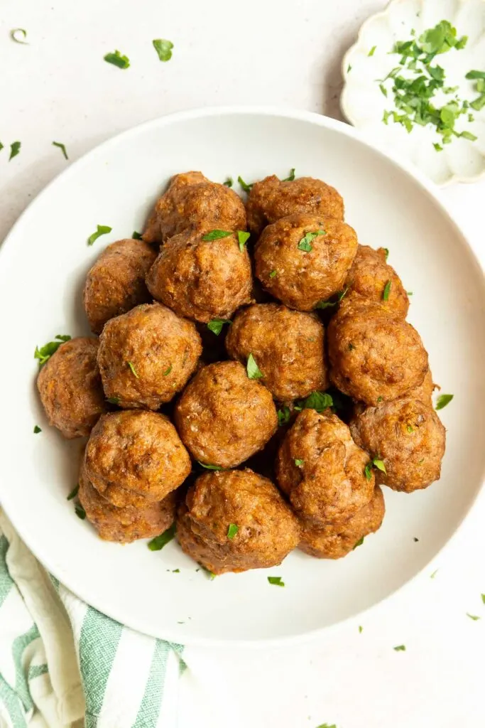A white bowl filled with browned Oven Baked Turkey Meatballs, garnished with chopped fresh parsley, sits on a light surface next to a small dish of more parsley and a striped kitchen towel.