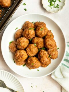 A white bowl filled with golden-brown baked ground turkey meatballs, garnished with chopped parsley, sits on a light surface next to a striped towel and a small dish of extra parsley.