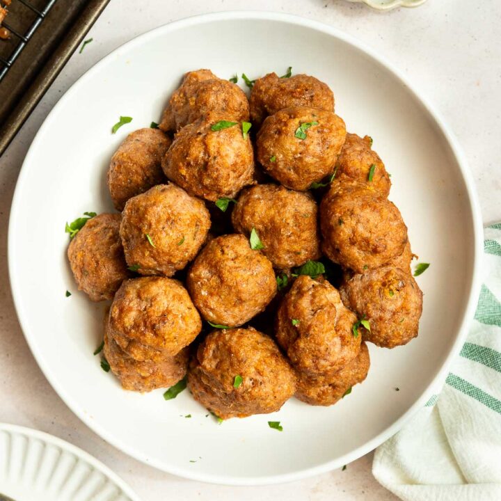 A white bowl filled with golden-brown baked ground turkey meatballs, garnished with chopped parsley, sits on a light surface next to a striped towel and a small dish of extra parsley.