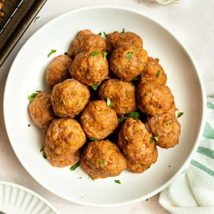 A white bowl filled with golden-brown baked ground turkey meatballs, garnished with chopped parsley, sits on a light surface next to a striped towel and a small dish of extra parsley.