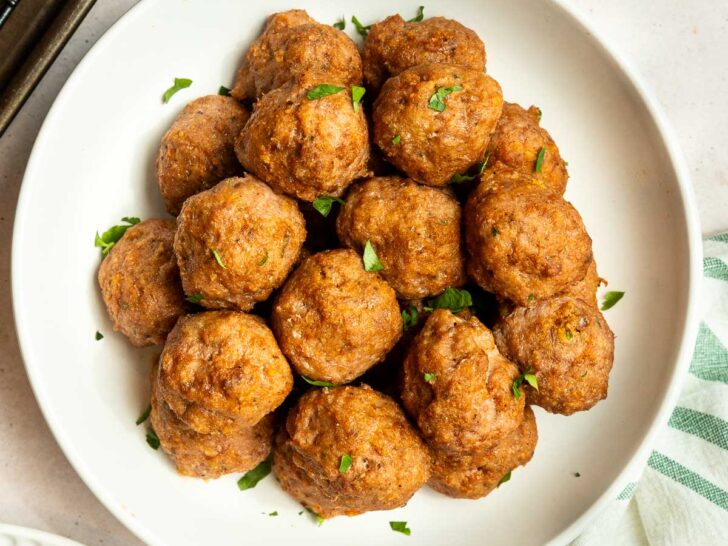 A white bowl filled with golden-brown baked ground turkey meatballs, garnished with chopped parsley, sits on a light surface next to a striped towel and a small dish of extra parsley.