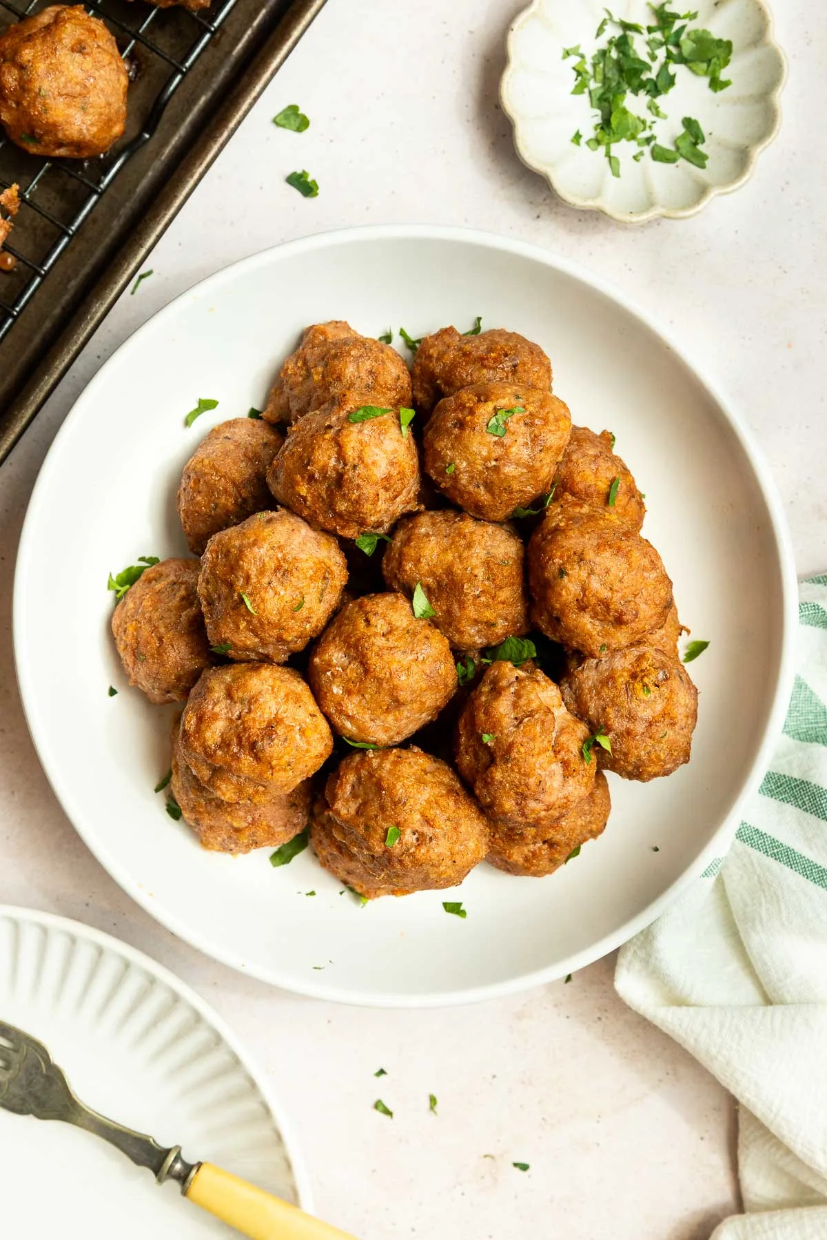 A white bowl filled with golden-brown baked ground turkey meatballs, garnished with chopped parsley, sits on a light surface next to a striped towel and a small dish of extra parsley.