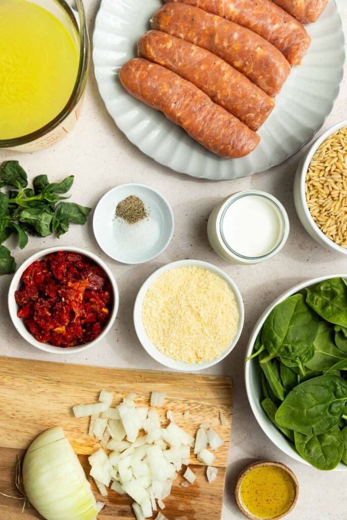 Overhead view of ingredients on a counter: italian sausage orzo on a plate, chopped onions, spinach, sun-dried tomatoes, grated cheese, milk, broth, seasonings, and fresh basil.