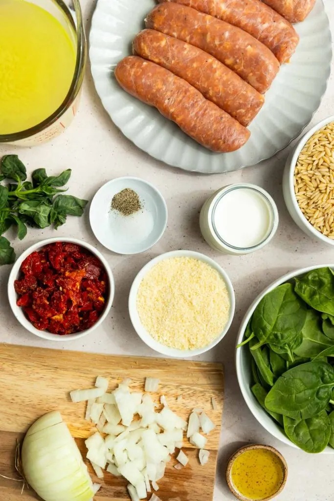 Overhead view of ingredients on a counter: italian sausage orzo on a plate, chopped onions, spinach, sun-dried tomatoes, grated cheese, milk, broth, seasonings, and fresh basil.