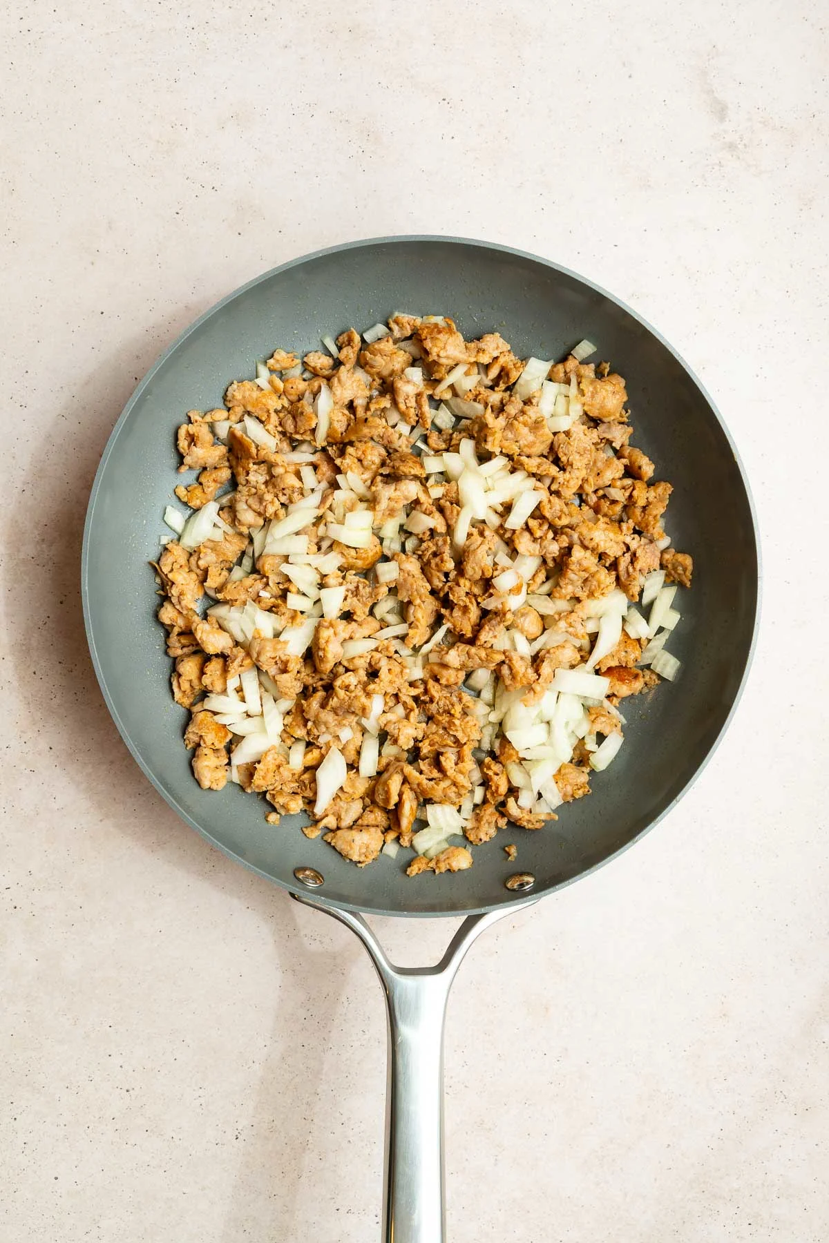 A nonstick skillet containing cooked ground meat and chopped onions, seen from above on a light-colored surface—perfect as a savory base for orzo with sausage.