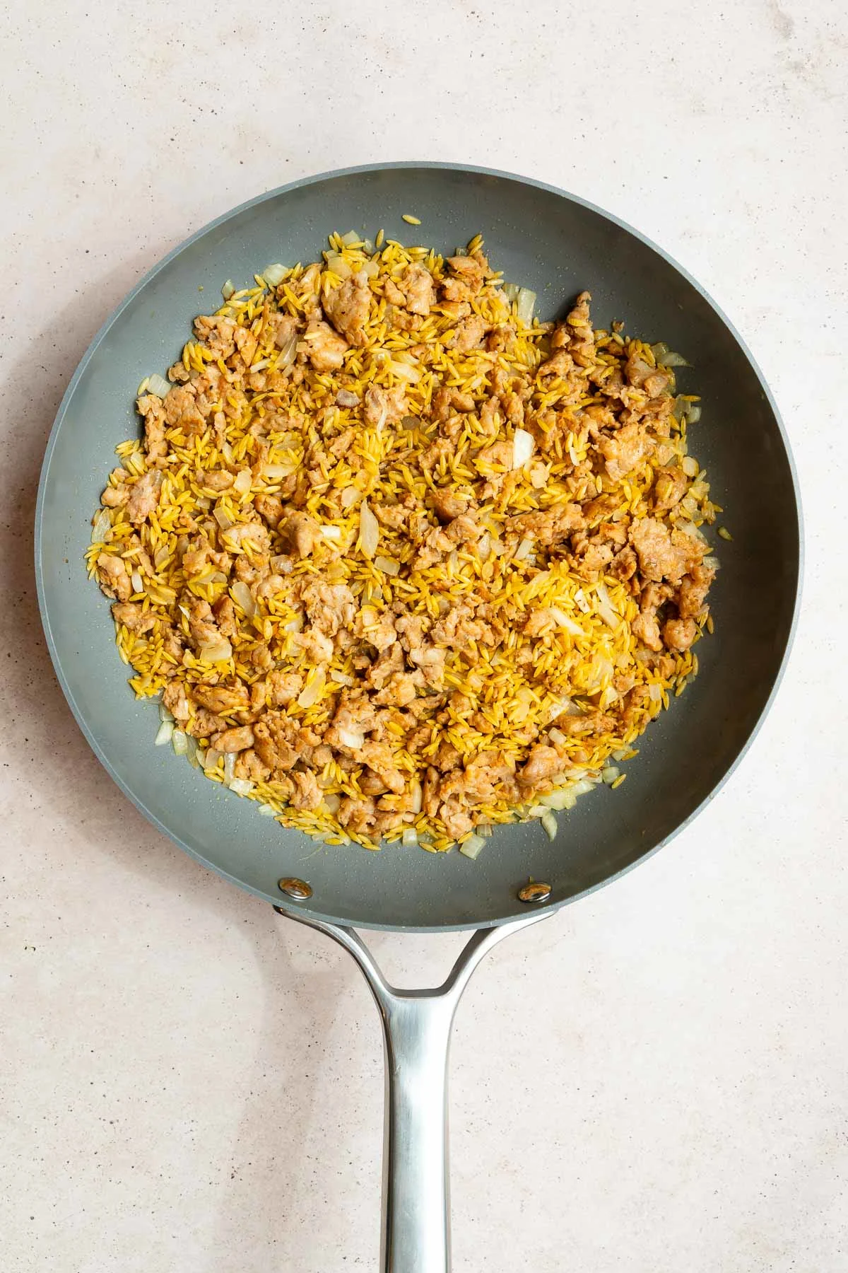 A gray frying pan filled with cooked yellow rice, ground meat, chopped onions, and savory sausage orzo pasta sits on a light-colored countertop.