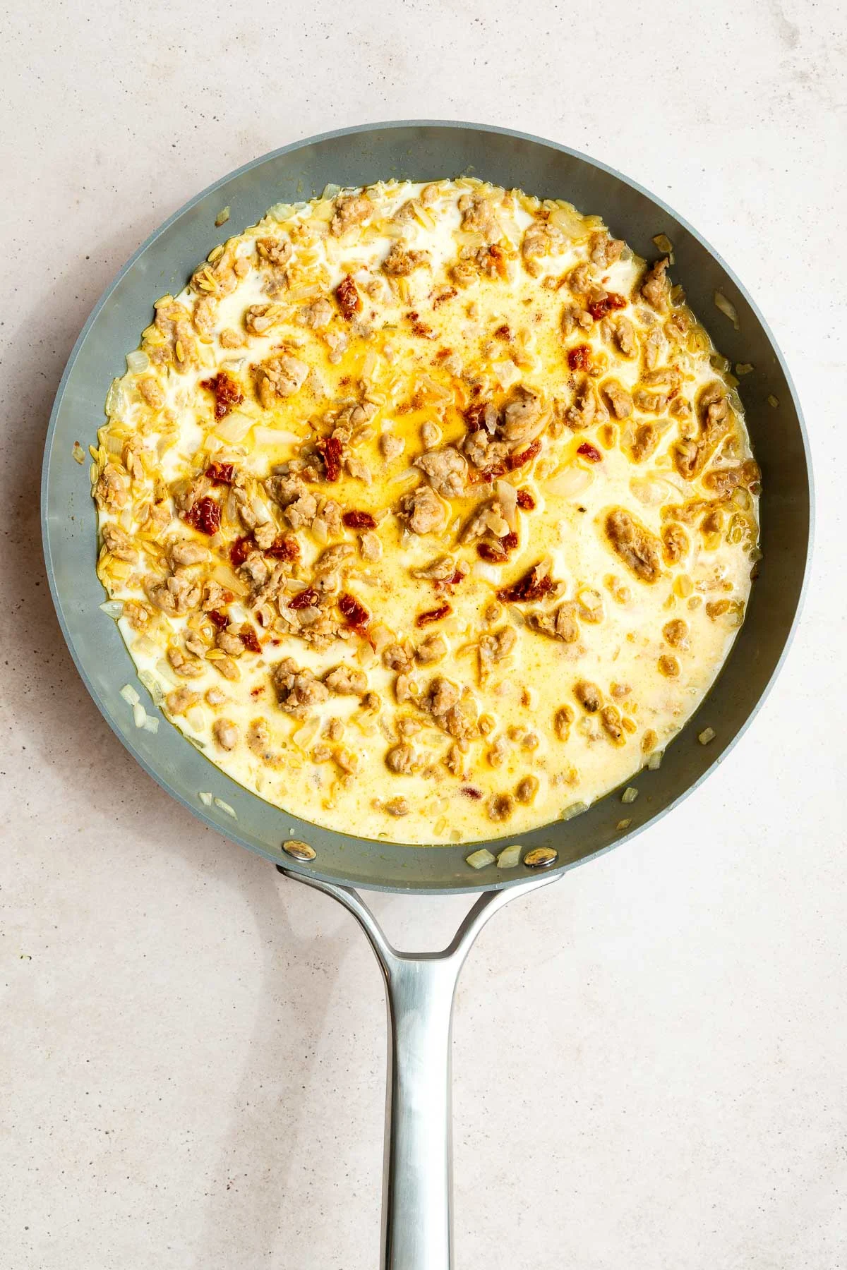 A skillet filled with a creamy one pot sausage orzo mixture and sun-dried tomatoes, creating a sauce with visible oil and spices, set against a light countertop.