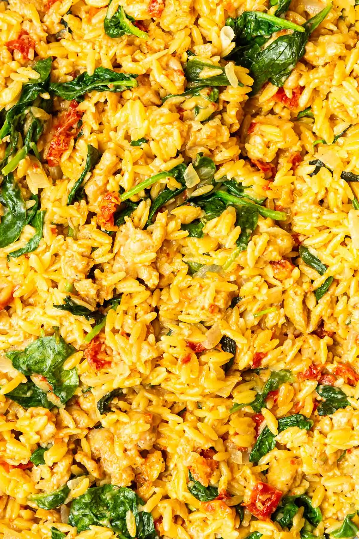 A close-up of cooked orzo pasta mixed with wilted spinach, sun-dried tomatoes, and small pieces of chicken in a creamy yellow sauce, reminiscent of a one pot creamy orzo with sausage and spinach.