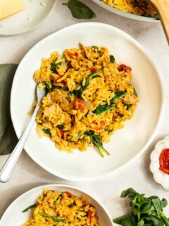 A white plate filled with creamy orzo pasta mixed with spinach, sun-dried tomatoes, and Italian sausage, garnished with herbs and a fork on the side. Other plates of this Creamy Italian Sausage Orzo Recipe and fresh basil are nearby.
