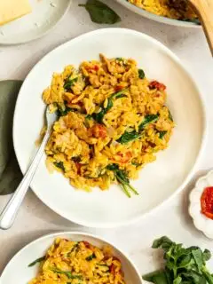 A white plate filled with creamy orzo pasta mixed with spinach, sun-dried tomatoes, and Italian sausage, garnished with herbs and a fork on the side. Other plates of this Creamy Italian Sausage Orzo Recipe and fresh basil are nearby.
