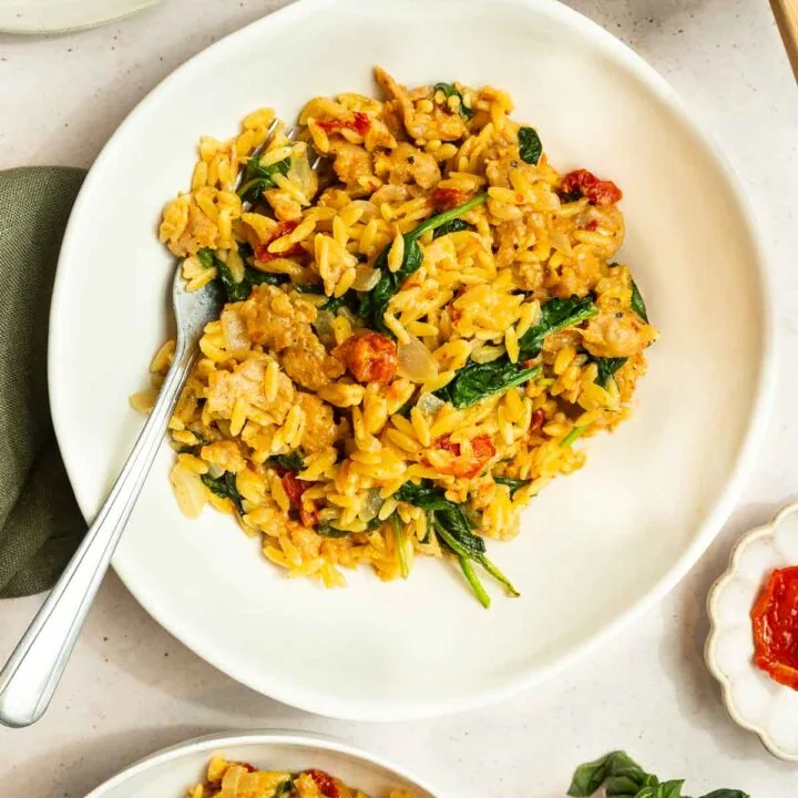 A white plate filled with creamy orzo pasta mixed with spinach, sun-dried tomatoes, and Italian sausage, garnished with herbs and a fork on the side. Other plates of this Creamy Italian Sausage Orzo Recipe and fresh basil are nearby.