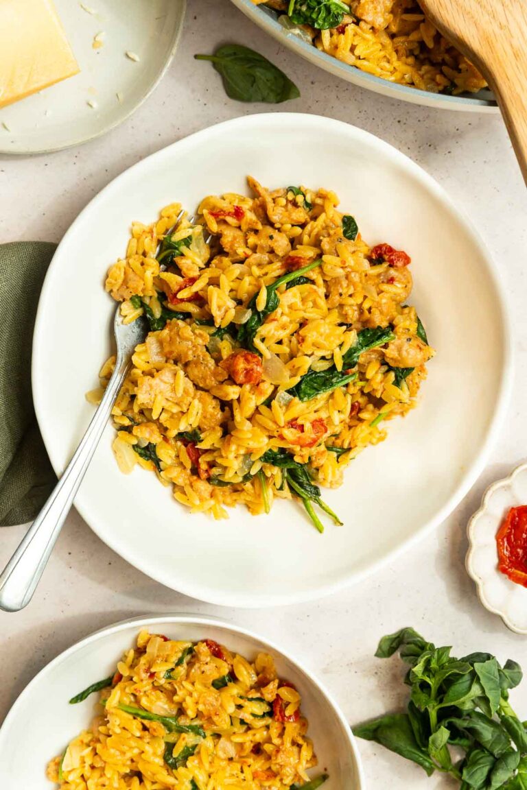 A white plate filled with creamy orzo pasta mixed with spinach, sun-dried tomatoes, and Italian sausage, garnished with herbs and a fork on the side. Other plates of this Creamy Italian Sausage Orzo Recipe and fresh basil are nearby.