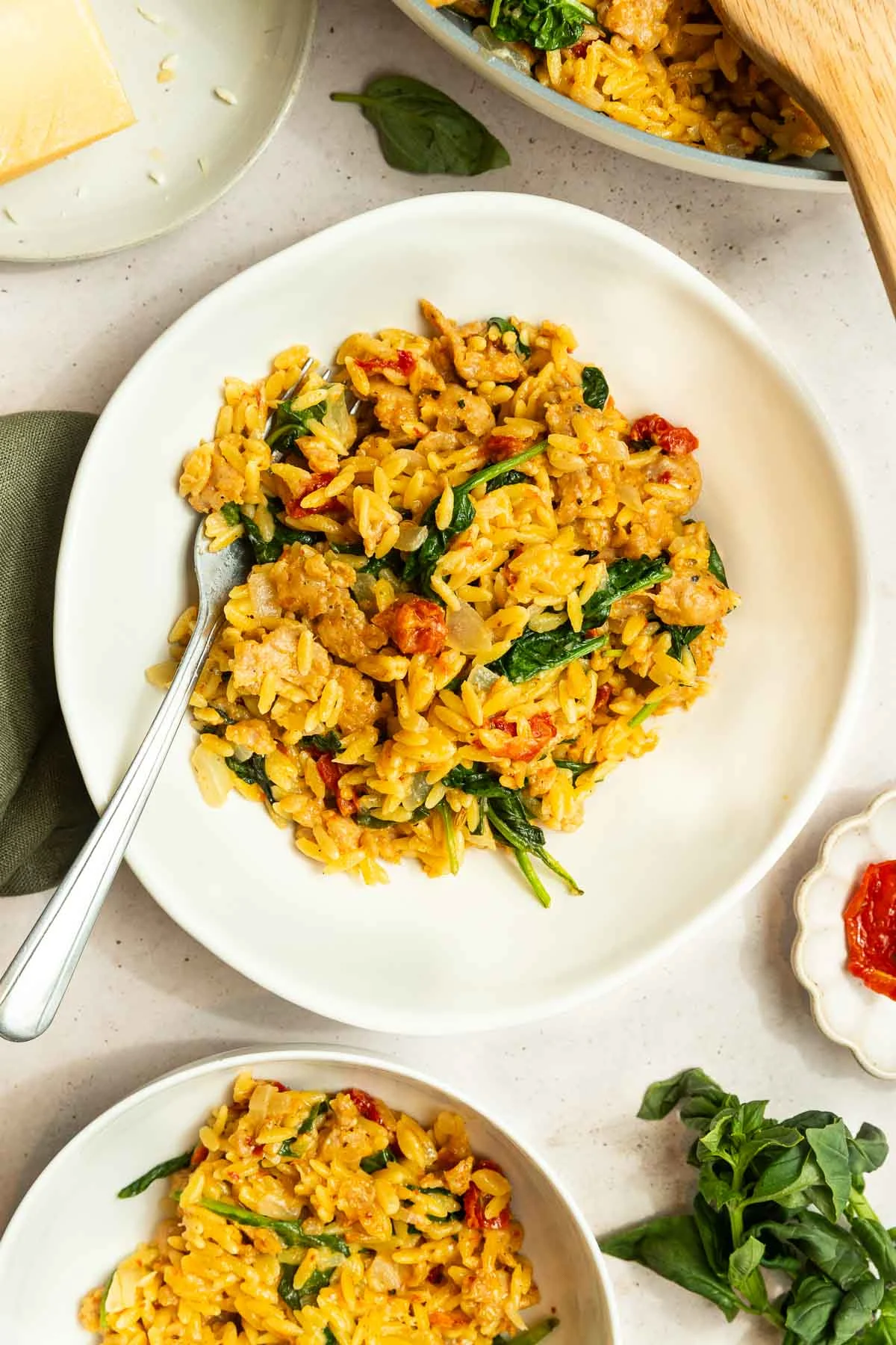 A white plate filled with creamy orzo pasta mixed with spinach, sun-dried tomatoes, and Italian sausage, garnished with herbs and a fork on the side. Other plates of this Creamy Italian Sausage Orzo Recipe and fresh basil are nearby.
