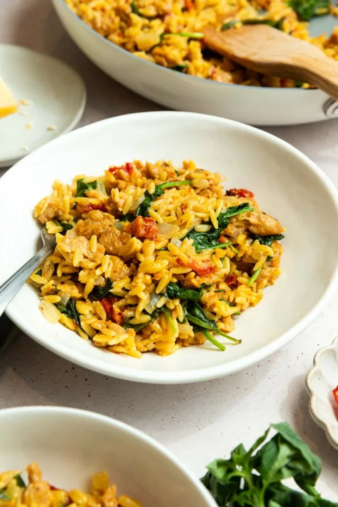 A white bowl filled with Italian sausage orzo pasta, mixed with spinach, sun-dried tomatoes, and pieces of chicken. A spoon rests in the bowl, with another serving of the same hearty dish partially visible in the background.
