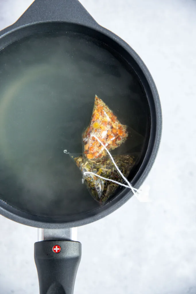 A black saucepan filled with hot water has two triangular tea bags steeping—one with orange and green herbs—creating a soothing medicine ball copycat brew. The pan rests on a light gray surface.
