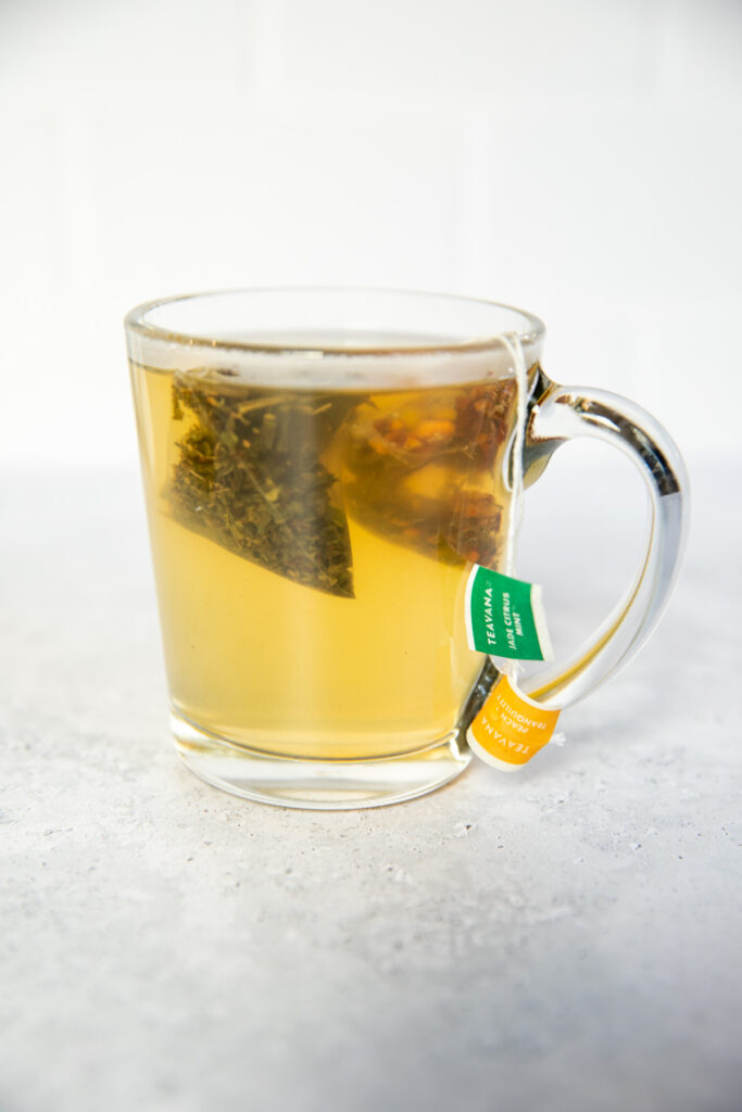 A glass mug filled with light herbal tea, resembling Starbucks Honey Citrus Mint Tea, with a tea bag inside and two tags—one green and one yellow—hanging over the side, set on a light, textured surface.