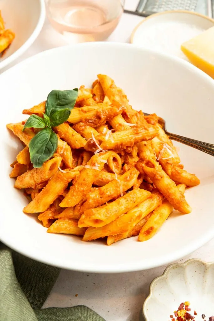 A white bowl filled with penne pasta coated in creamy vodka sauce, garnished with fresh basil leaves. A fork rests in the bowl, and a glass of rosé wine and a wedge of cheese are nearby.
