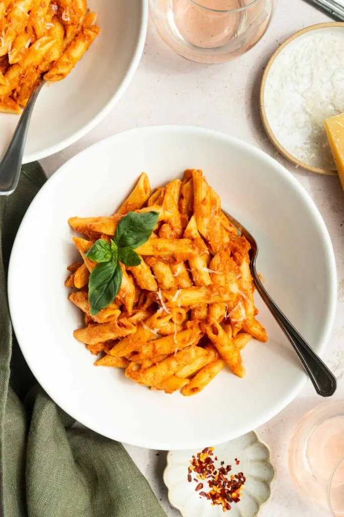 A white bowl of penne pasta with creamy spicy vodka sauce recipe, garnished with fresh basil, sits on a table with a fork. Nearby are a bowl of grated cheese, a glass of water, and a small dish of red pepper flakes.