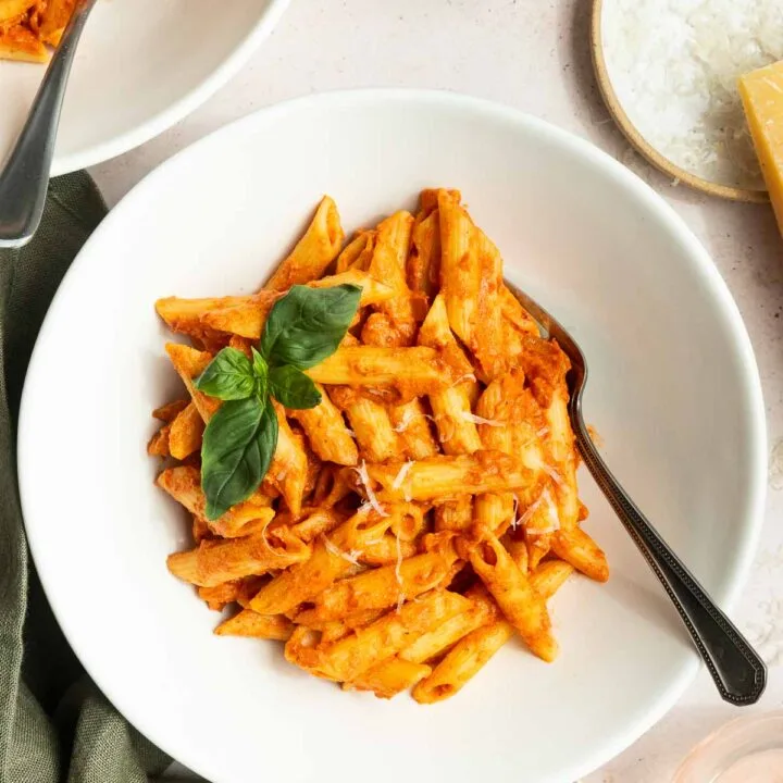 A white bowl of penne pasta with creamy spicy vodka sauce recipe, garnished with fresh basil, sits on a table with a fork. Nearby are a bowl of grated cheese, a glass of water, and a small dish of red pepper flakes.