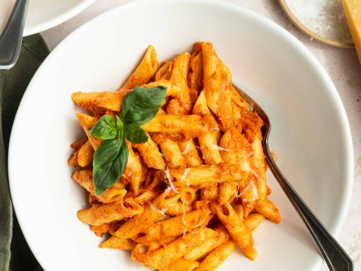 A white bowl of penne pasta with creamy spicy vodka sauce recipe, garnished with fresh basil, sits on a table with a fork. Nearby are a bowl of grated cheese, a glass of water, and a small dish of red pepper flakes.