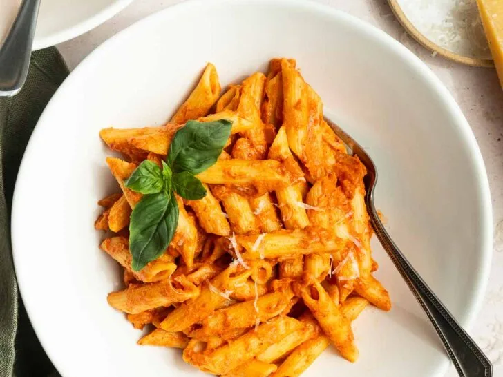 A white bowl of penne pasta with creamy spicy vodka sauce recipe, garnished with fresh basil, sits on a table with a fork. Nearby are a bowl of grated cheese, a glass of water, and a small dish of red pepper flakes.