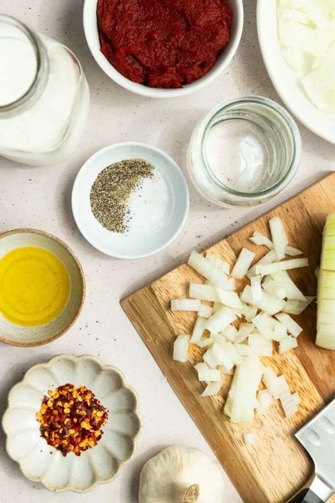 Overhead view of chopped onions on a cutting board, surrounded by bowls of tomato paste, diced onions, olive oil, milk, salt, pepper, red pepper flakes, and a garlic bulb—perfect ingredients for a classic vodka sauce recipe.